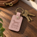 Blush Doxie Mom keychain on a dark walnut table beside brass keys, with a coiled leather leash in the background