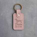 Blush pink leather keychain debossed with a dachshund silhouette and the words Doxie MOM, lying on gray concrete