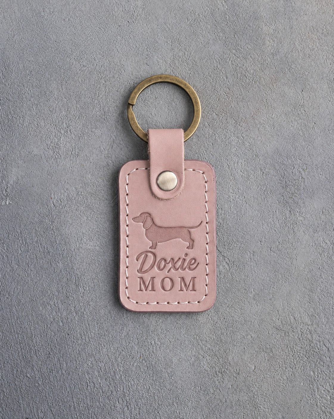 Blush pink leather keychain debossed with a dachshund silhouette and the words Doxie MOM, lying on gray concrete