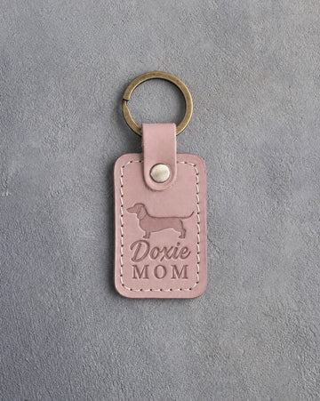 Blush pink leather keychain debossed with a dachshund silhouette and the words Doxie MOM, lying on gray concrete