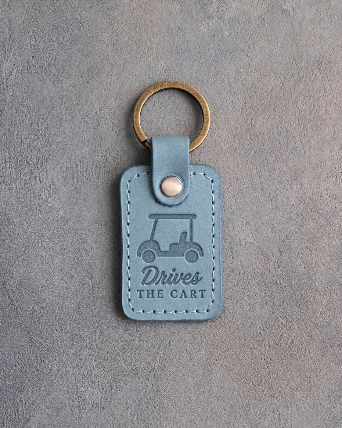 Turquoise leather keychain debossed with a golf cart above the words Drives THE CART, on gray plaster