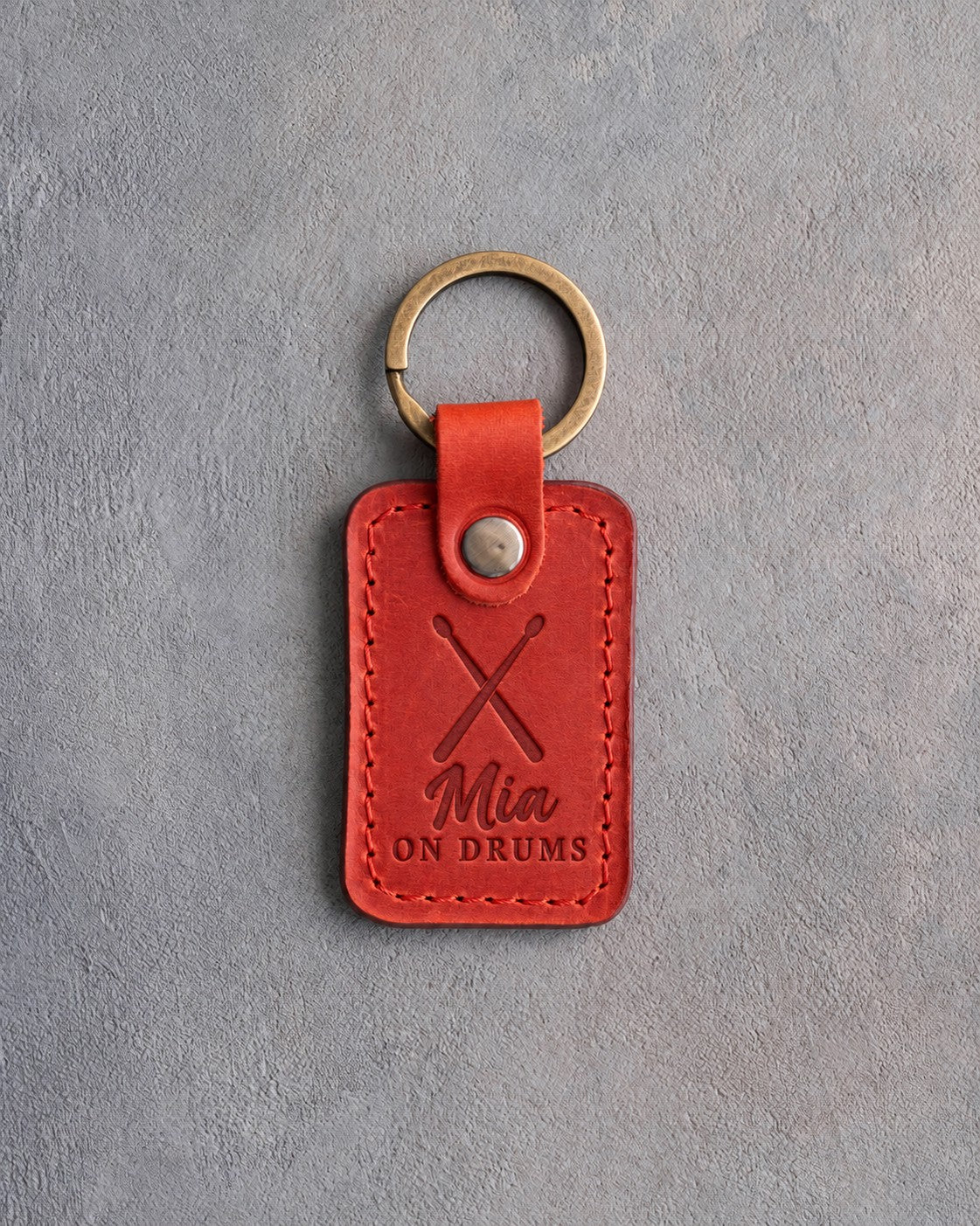 Poppy red leather keychain debossed with crossed drumsticks above the name Mia and ON DRUMS, on gray plaster