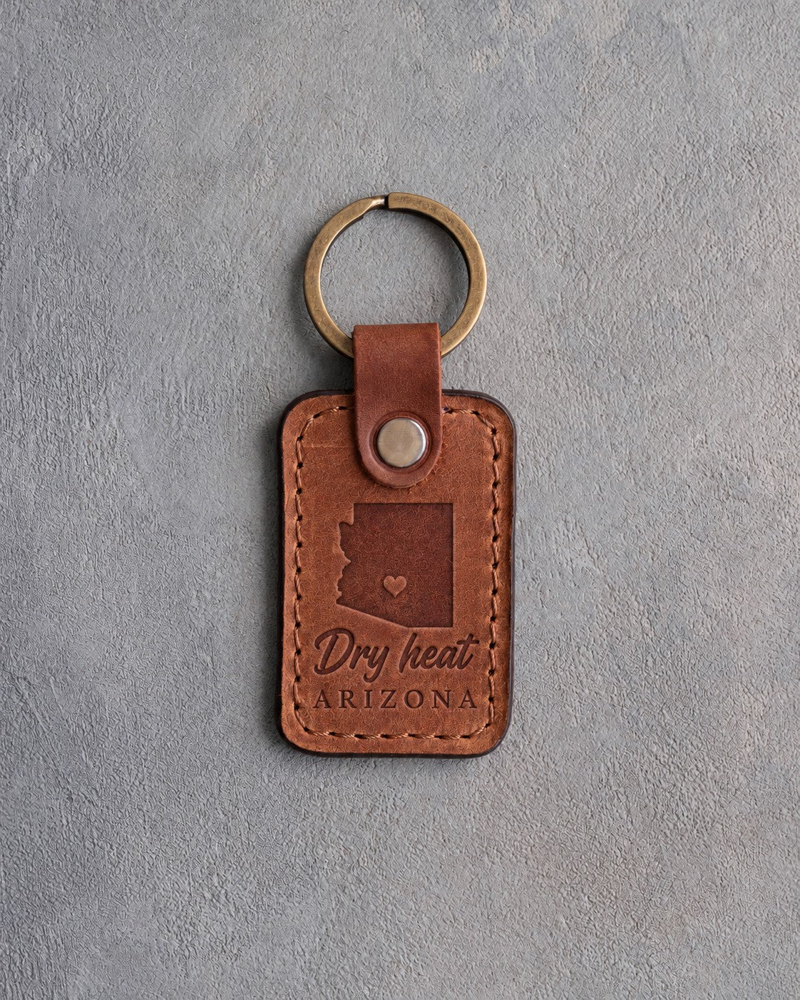 Sandy tan leather keychain debossed with a solid Arizona silhouette and a heart at Phoenix above Dry heat and ARIZONA, on gray concrete