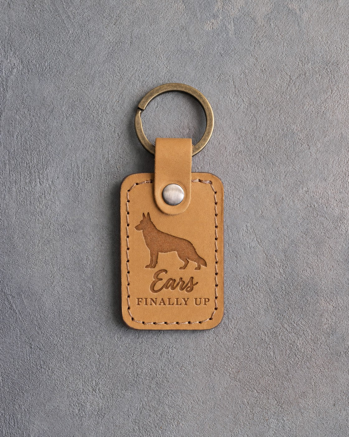 Indian Summer tan leather keychain debossed with a German shepherd silhouette with upright ears and the words Ears FINALLY UP, on gray concrete