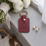 Engaged DEC 24 2026 keychain in wine red leather on a gray windowsill beside pearl earrings and a linen-wrapped bouquet