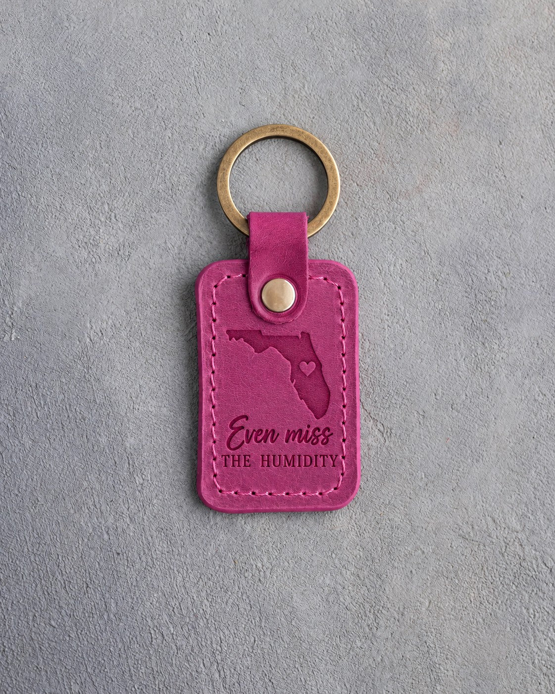 Fuchsia pink leather keychain debossed with a Florida silhouette and a heart at Orlando above Even miss THE HUMIDITY, on gray concrete