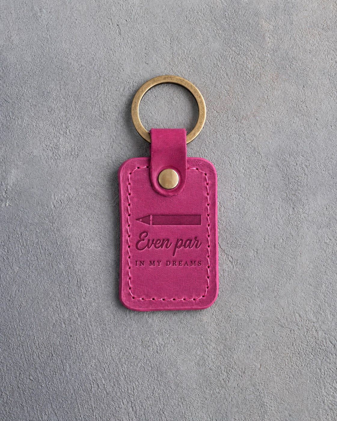 Fuchsia pink leather keychain debossed with a scorecard pencil above Even par IN MY DREAMS, on gray plaster