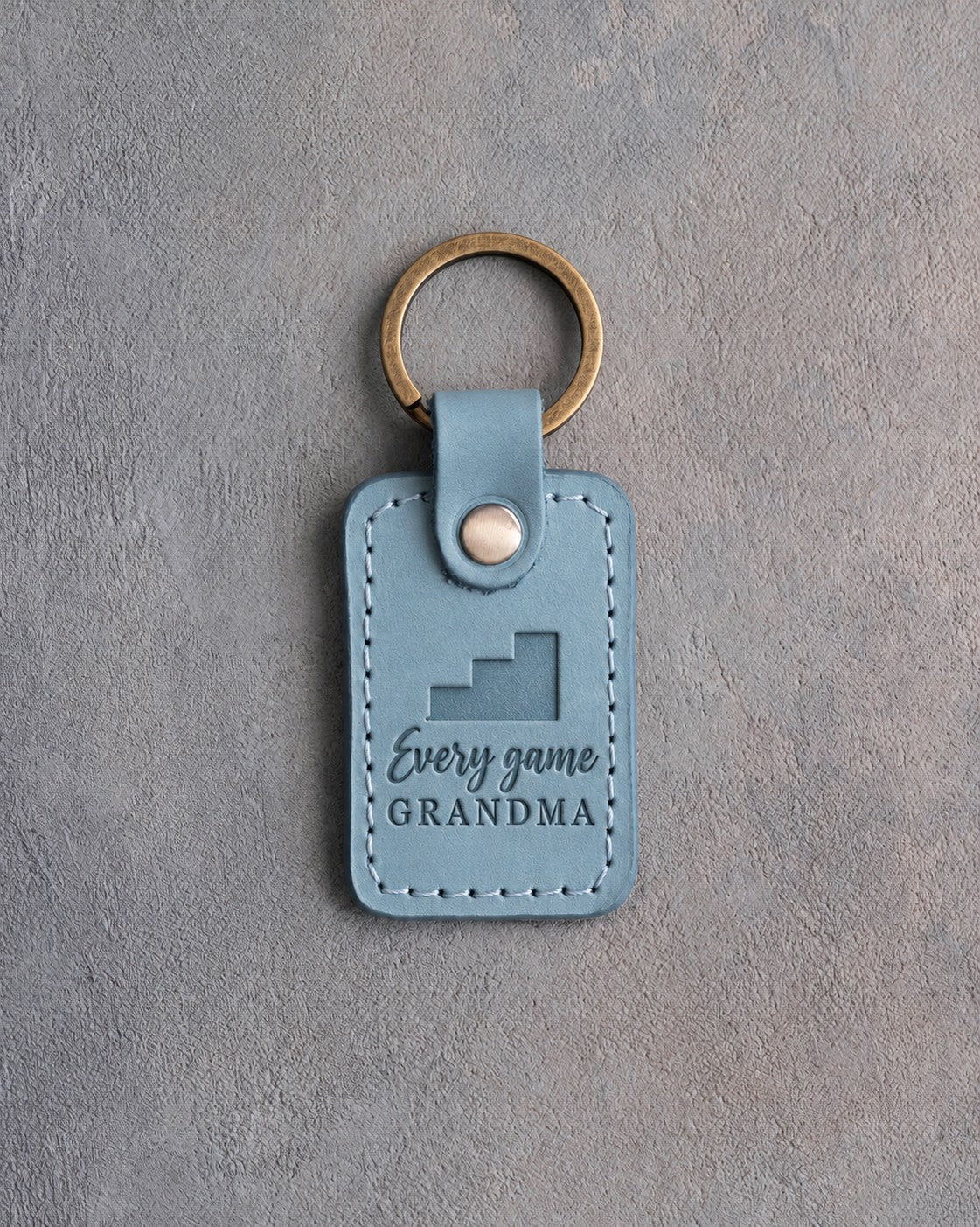 Turquoise leather keychain debossed with stepped bleachers and the words Every game GRANDMA, on gray plaster