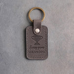 Country gray leather keychain debossed with a trophy cup and the words Every game GRANDPA, on gray plaster
