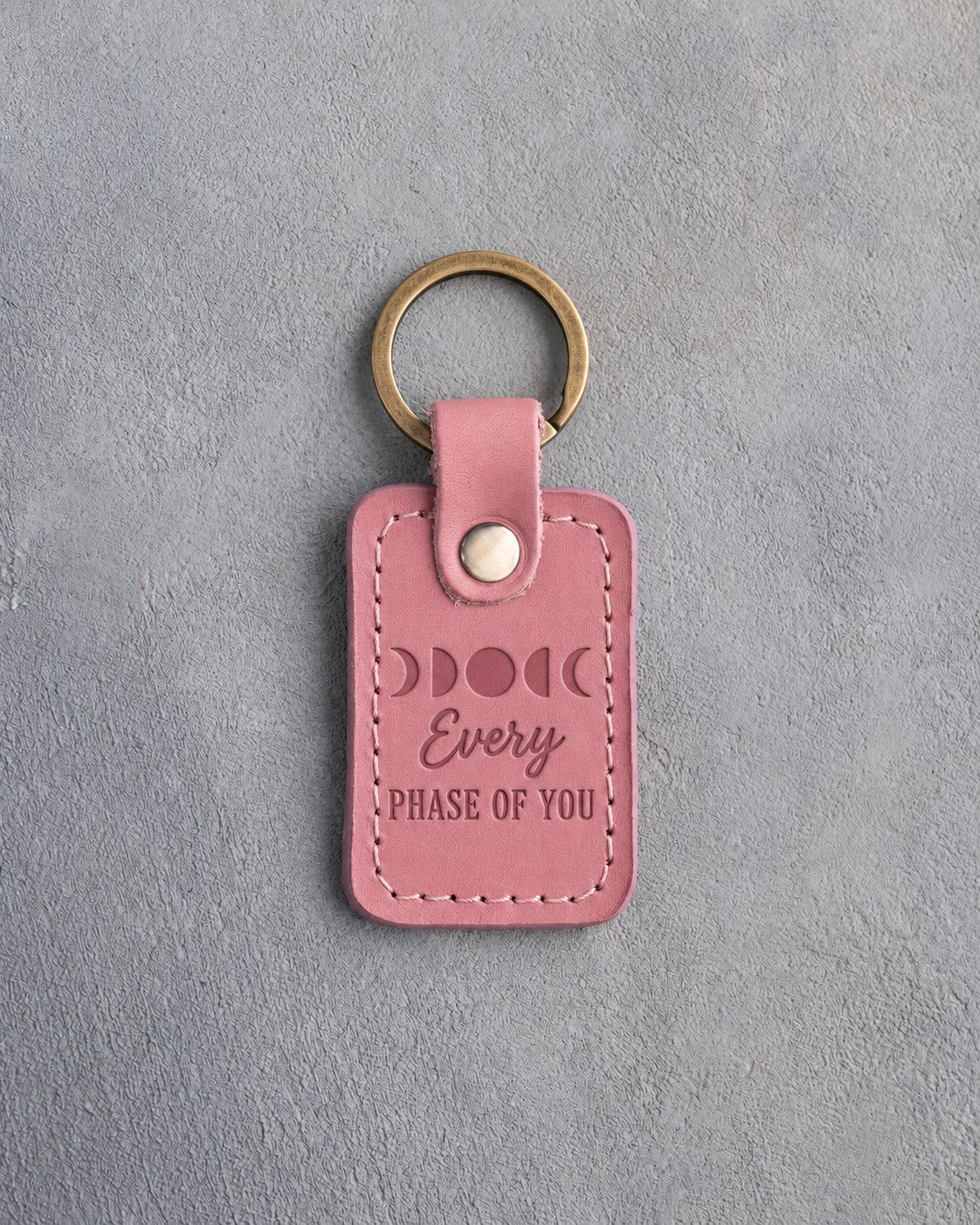 Rose pink leather keychain debossed with a row of five moon phases above Every and PHASE OF YOU, on gray concrete