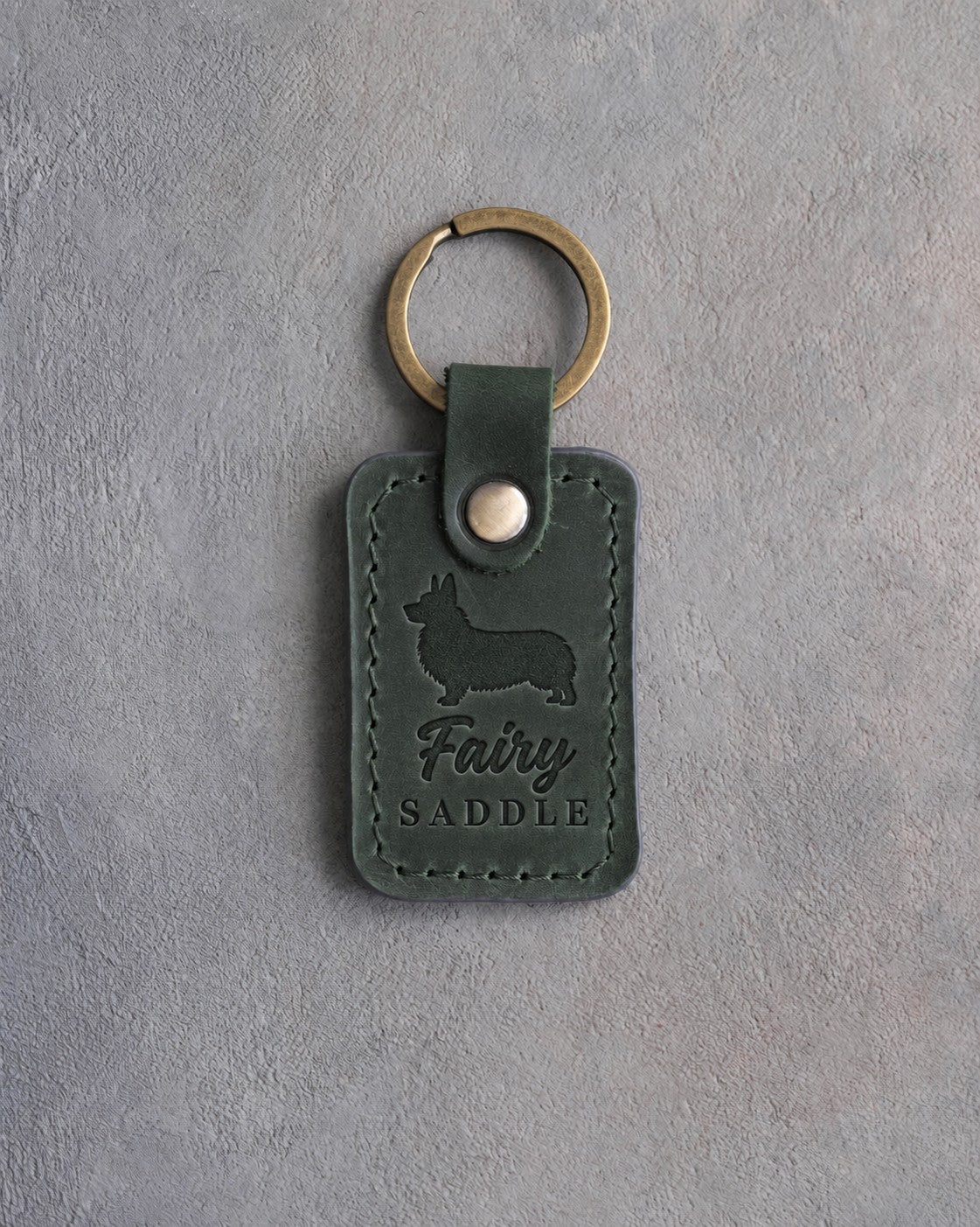 Forest green leather keychain debossed with a Pembroke corgi silhouette and the words Fairy SADDLE, on gray concrete