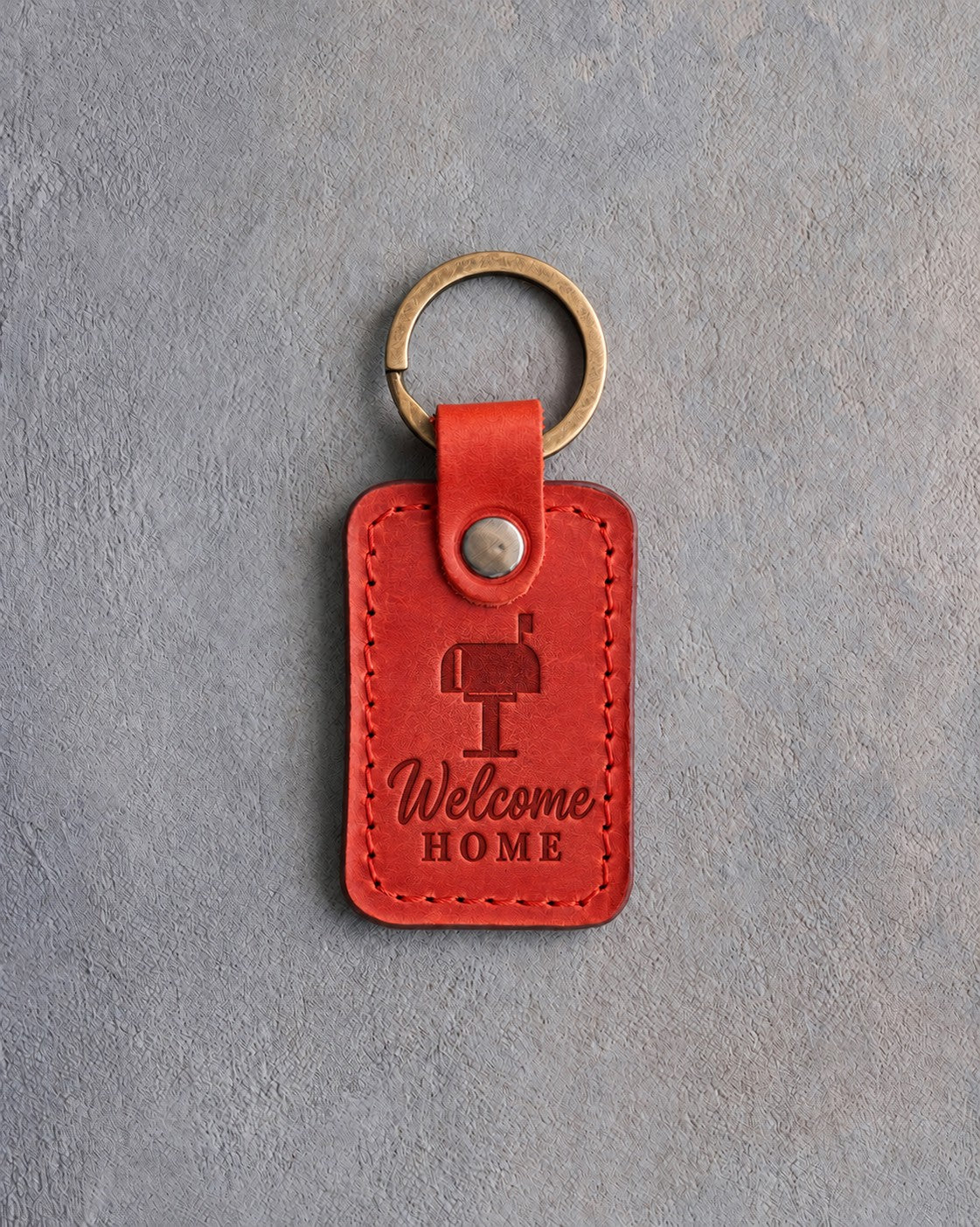 Poppy red leather keychain debossed with a roadside mailbox above Welcome and HOME, on gray concrete