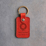 Poppy red leather keychain debossed with a Christmas wreath above Merry Christmas and DECEMBER 2026, on gray concrete