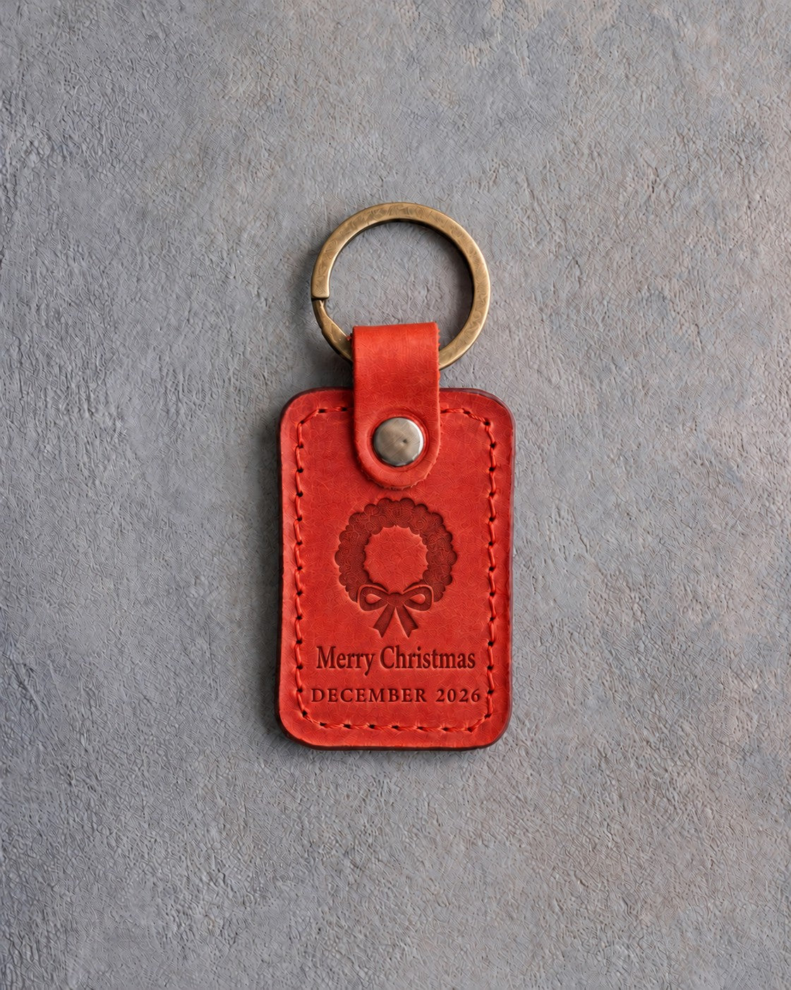 Poppy red leather keychain debossed with a Christmas wreath above Merry Christmas and DECEMBER 2026, on gray concrete