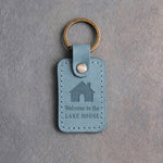Turquoise leather keychain debossed with a gabled house above Welcome to the and LAKE HOUSE, on gray concrete