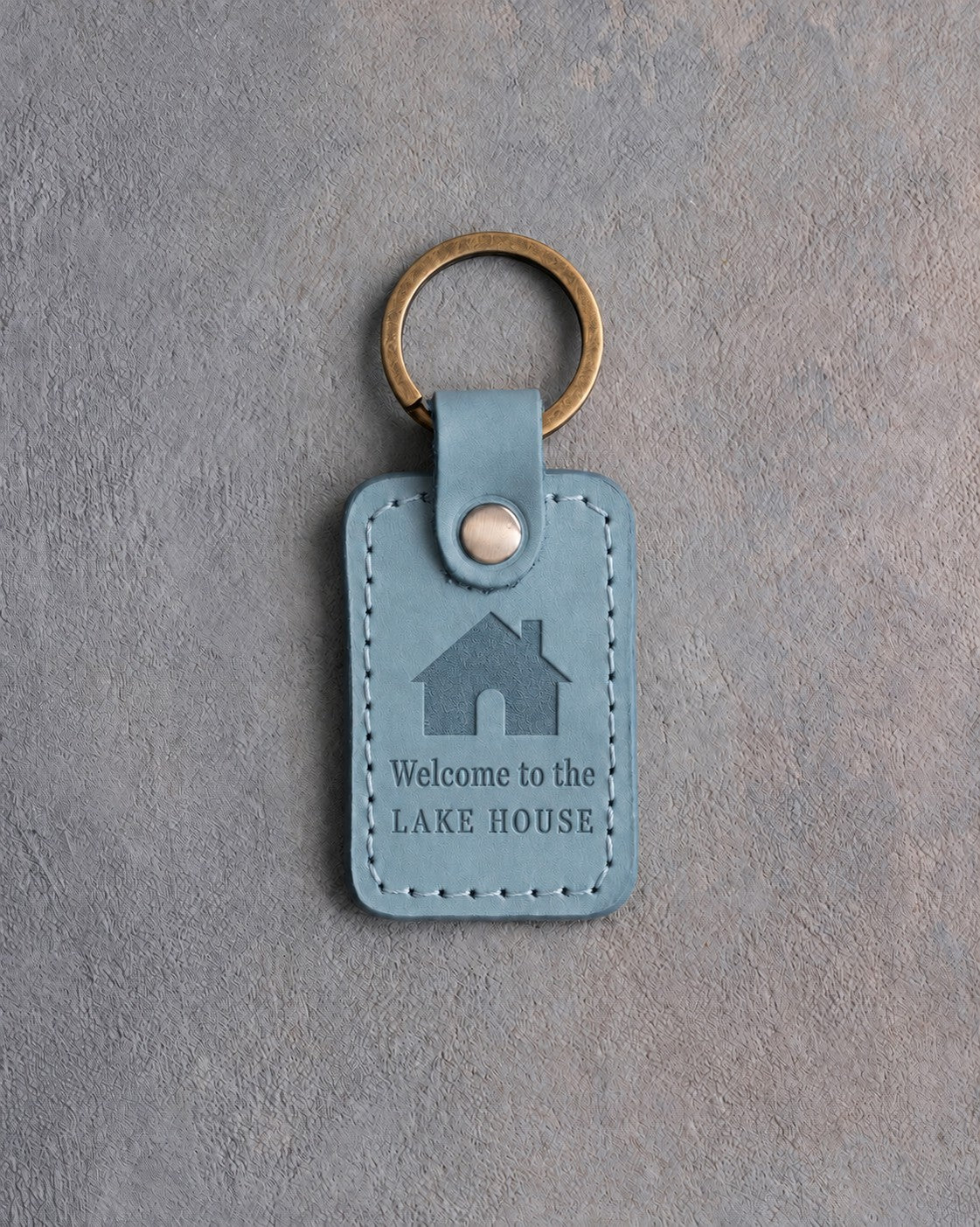 Turquoise leather keychain debossed with a gabled house above Welcome to the and LAKE HOUSE, on gray concrete