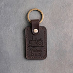 Dark espresso leather keychain debossed with an open-station tractor above the words Farm DAD, on gray concrete