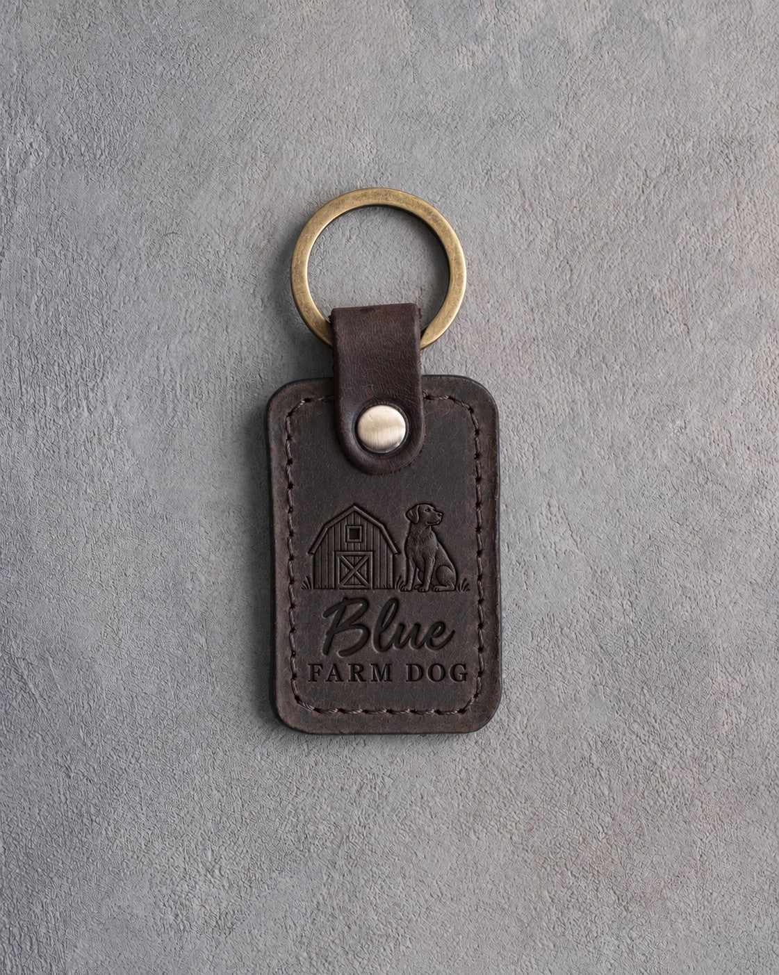 Dark espresso leather keychain debossed with a barn and a dog above Blue and FARM DOG, on gray concrete