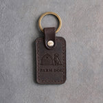 Dark espresso leather keychain debossed with a barn and a seated dog above FARM DOG, on gray concrete
