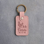 Blush pink leather keychain debossed with a hen and two chicks above the words Farm MOM, on gray concrete