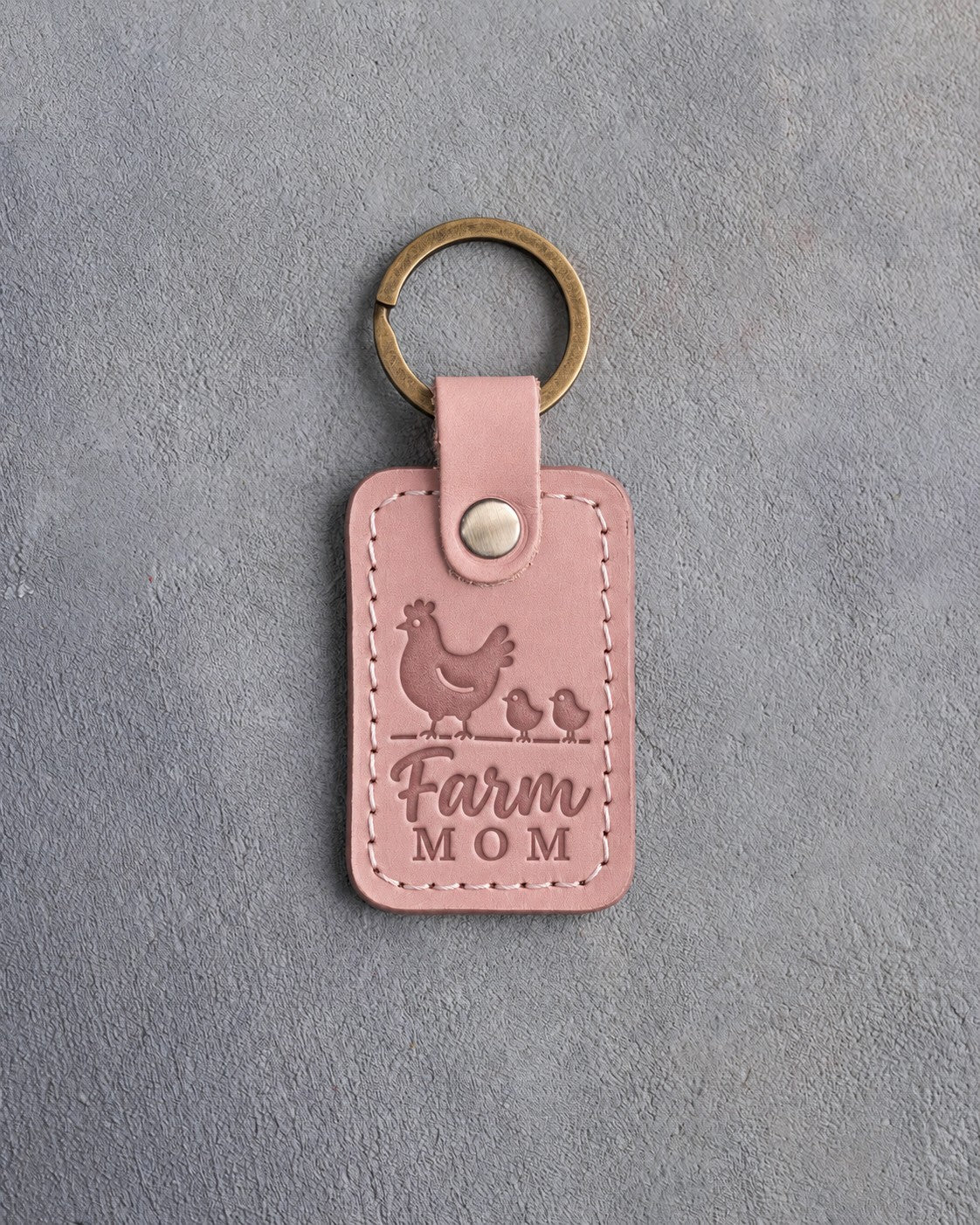 Blush pink leather keychain debossed with a hen and two chicks above the words Farm MOM, on gray concrete