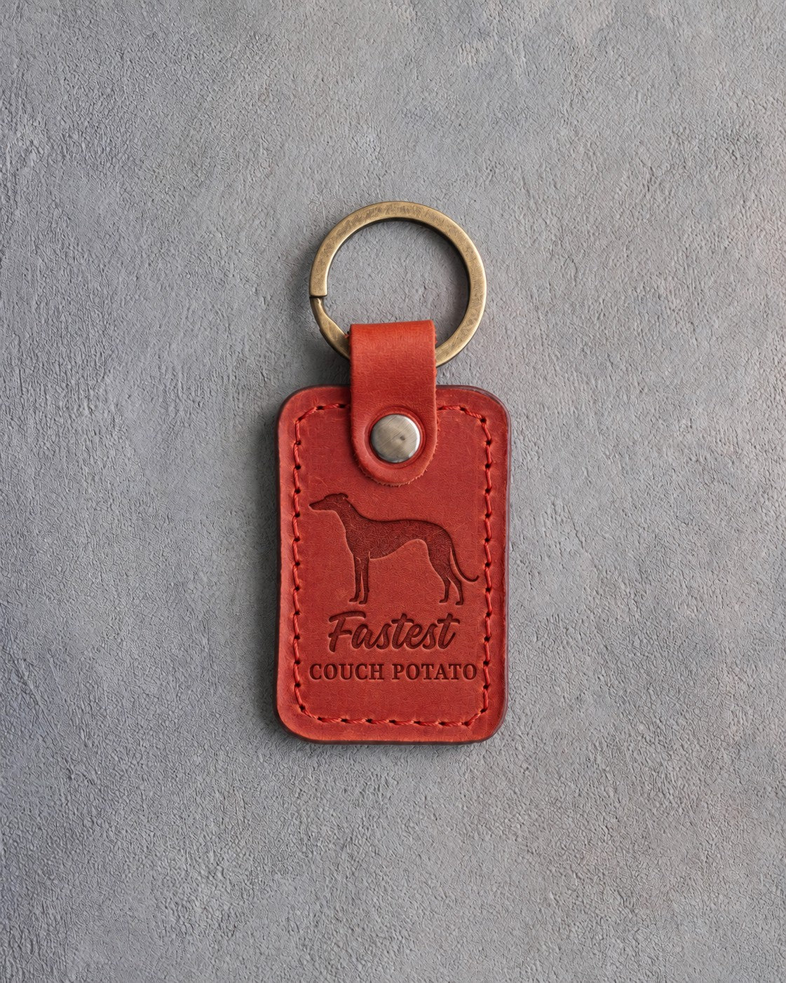 Poppy red leather keychain debossed with a greyhound and the words Fastest COUCH POTATO, on gray concrete