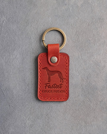 Poppy red leather keychain debossed with a greyhound and the words Fastest COUCH POTATO, on gray concrete