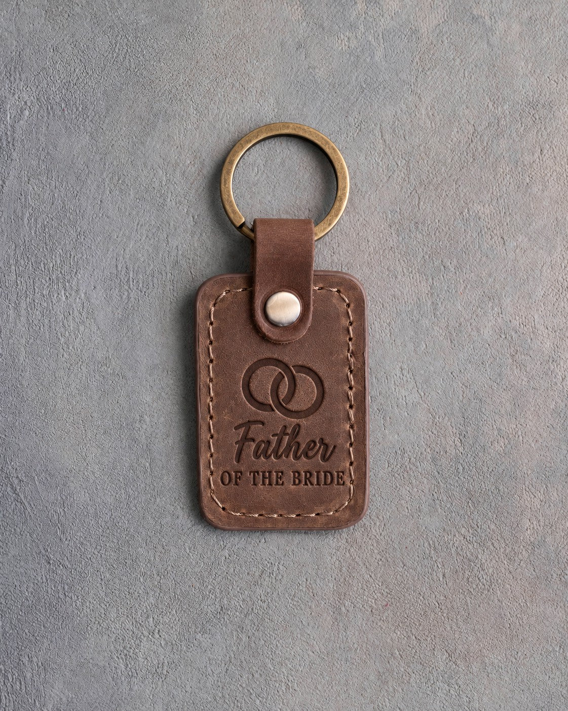 Sicilian brown leather keychain debossed with two interlocked wedding rings above Father and OF THE BRIDE, on gray concrete