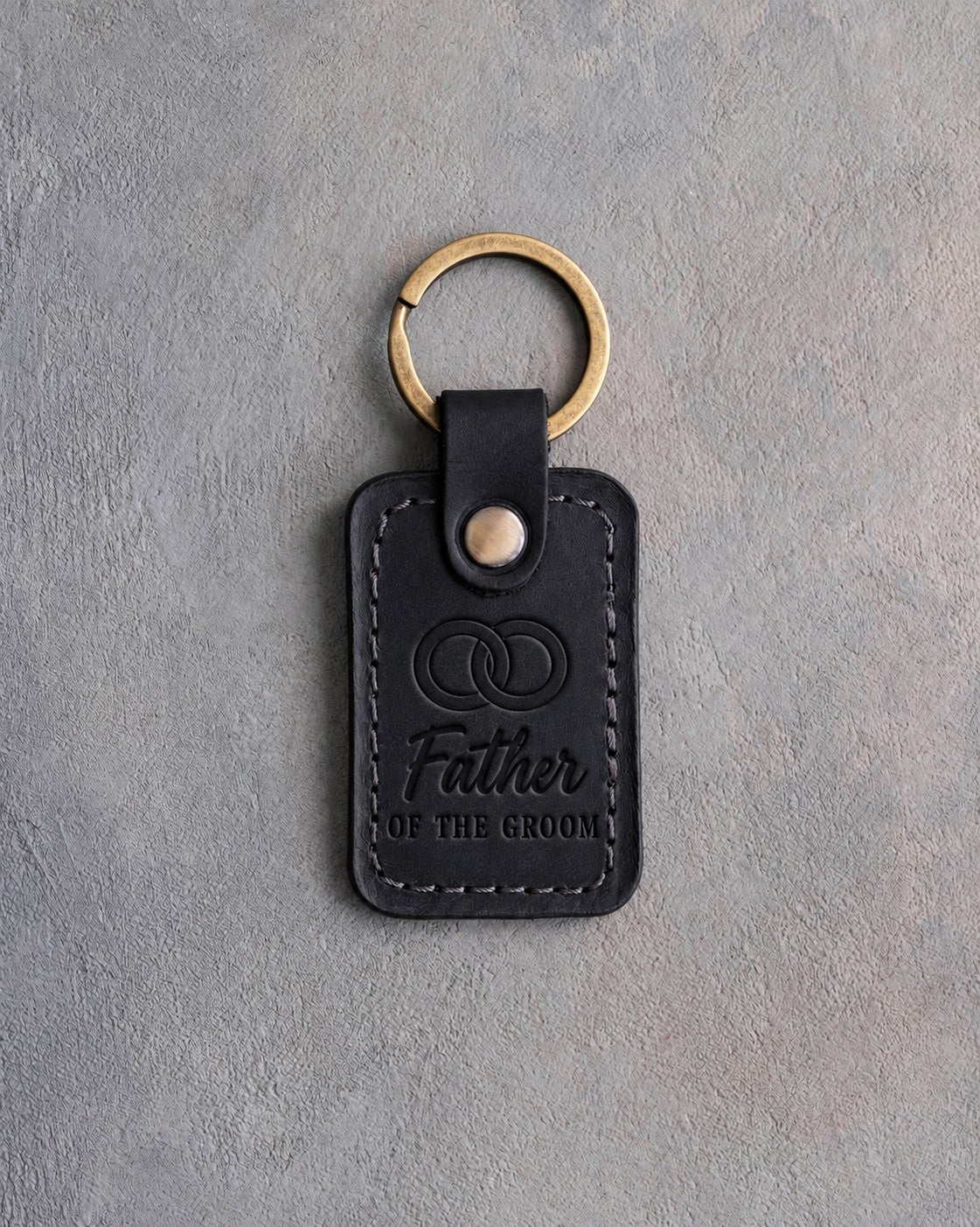 Black leather keychain debossed with two interlocked wedding rings above Father and OF THE GROOM, on gray concrete