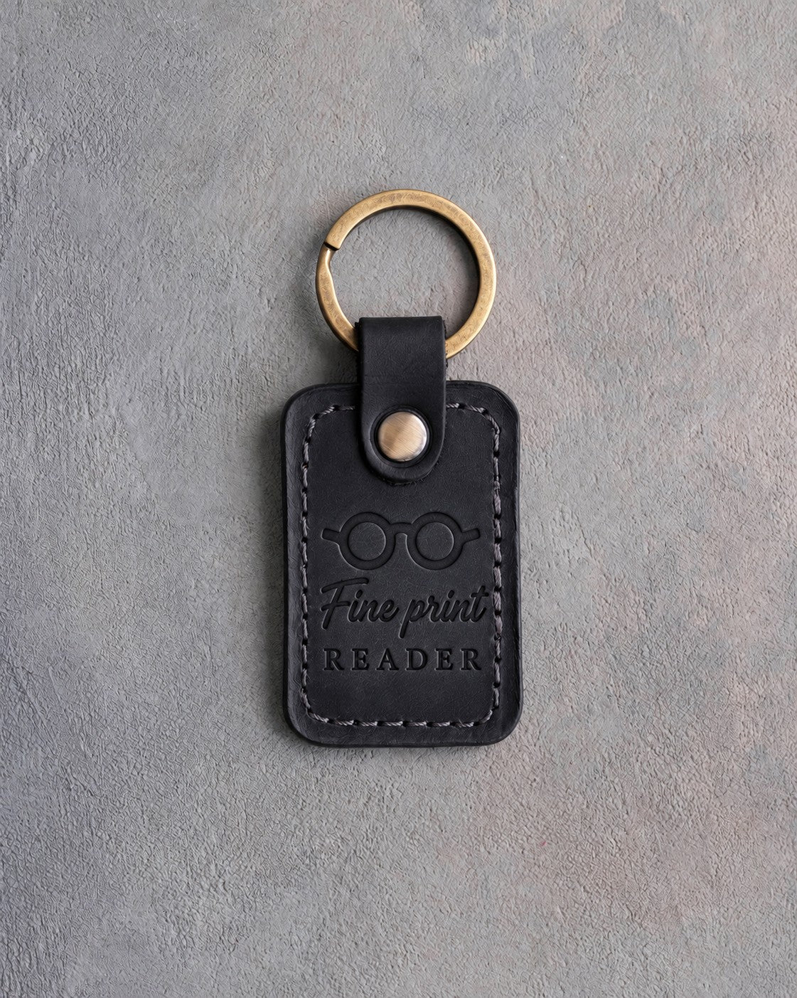 Black leather keychain debossed with round reading glasses above Fine print and READER, on gray concrete