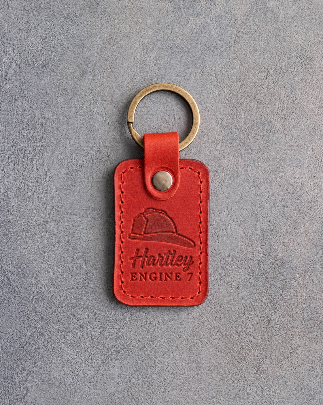 Red leather keychain debossed with a side-profile fire helmet above the name Hartley and ENGINE 7, on gray plaster