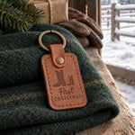 Caramel First CHRISTMAS keychain on a folded green wool blanket inside a barn door, a kraft parcel tied with twine behind it and snow outside