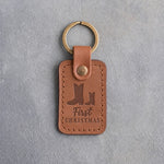 Caramel leather keychain debossed with a big and a small cowboy boot above First and CHRISTMAS, on gray plaster