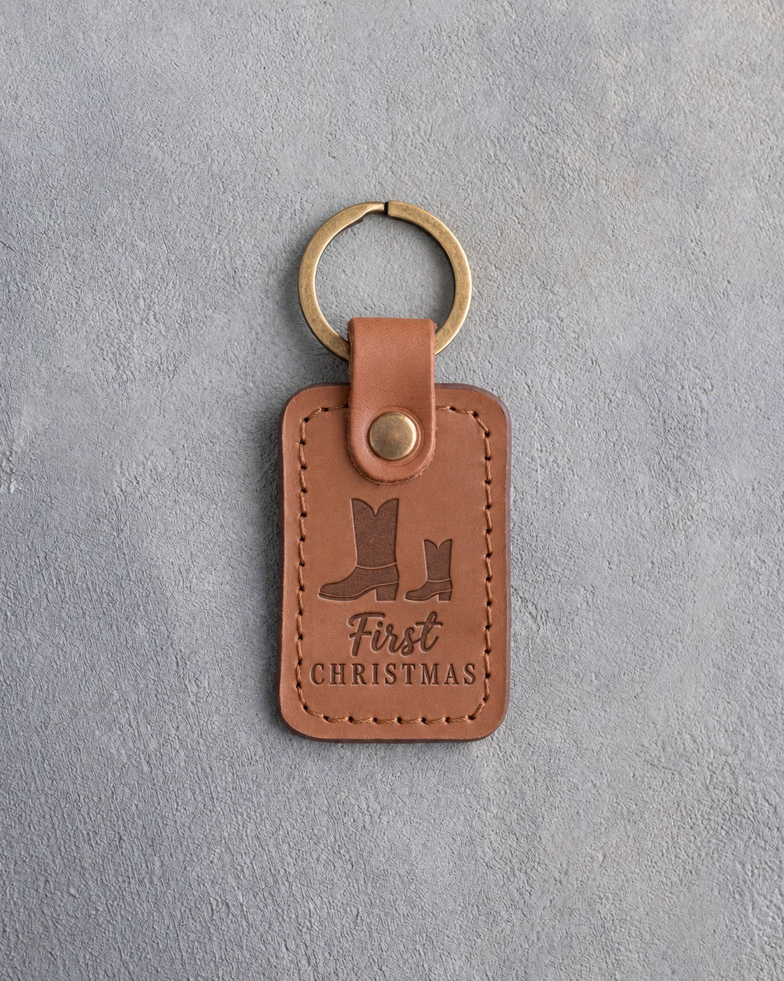Caramel leather keychain debossed with a big and a small cowboy boot above First and CHRISTMAS, on gray plaster