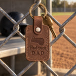 Brown First Coach Dad keychain hanging on a chain-link dugout fence in warm late-afternoon light
