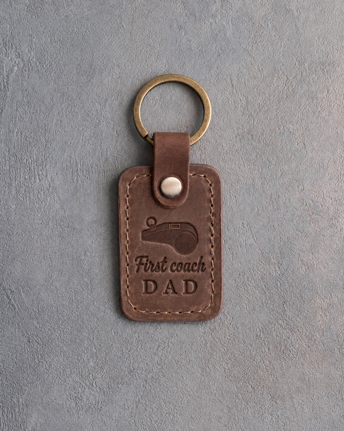 Sicilian brown leather keychain debossed with a coach's whistle and the words First coach DAD, on gray plaster