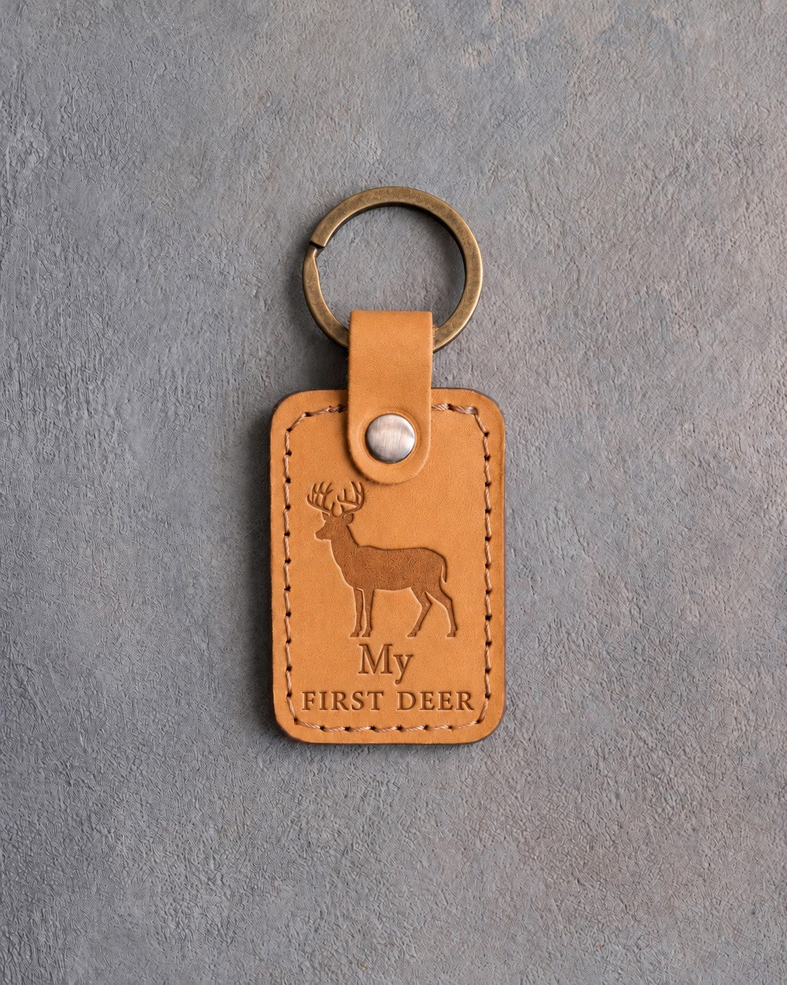 Golden tan leather keychain debossed with a whitetail buck above My and FIRST DEER, on gray concrete