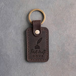 Dark espresso leather keychain debossed with a quill in an inkwell above First draft and DONE, on gray concrete