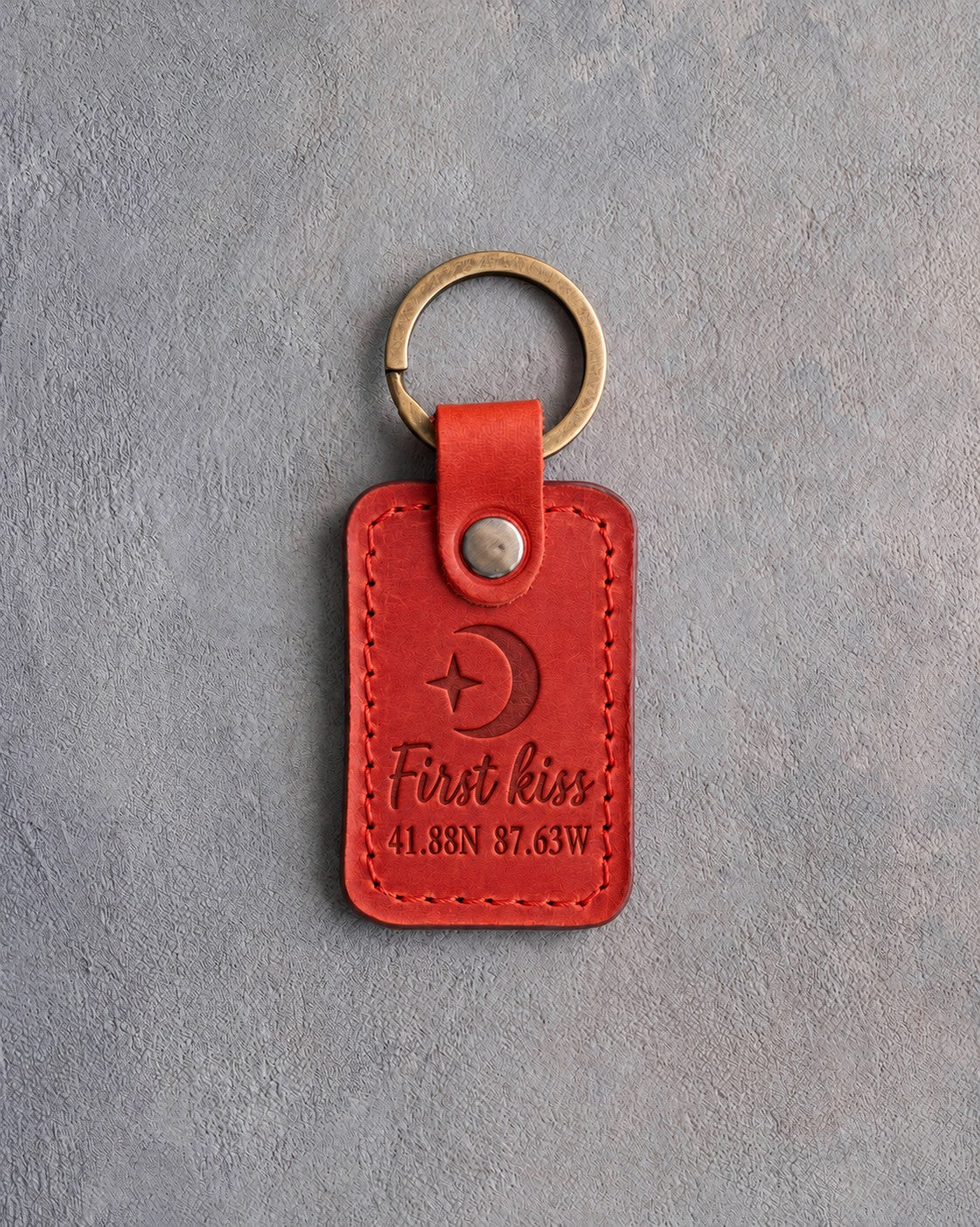 Poppy red leather keychain debossed with a crescent moon and star above First kiss and 41.88N 87.63W, on gray concrete