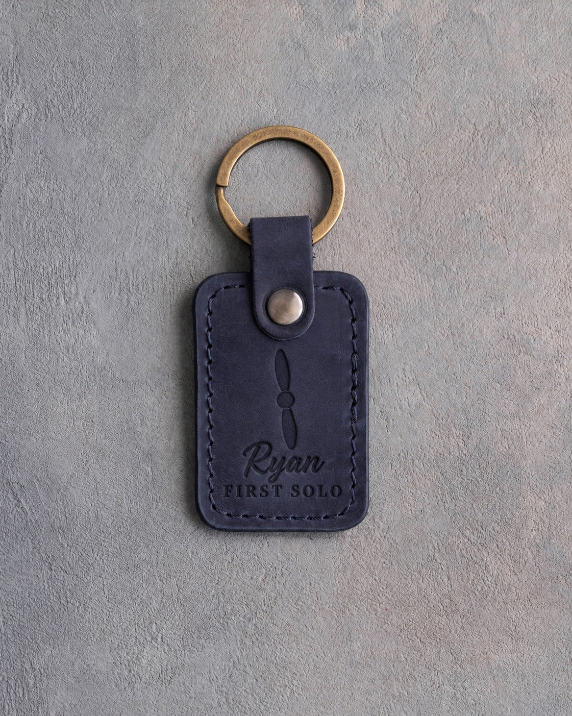 Navy blueberry leather keychain debossed with a two-blade propeller above the name Ryan and FIRST SOLO, on gray plaster