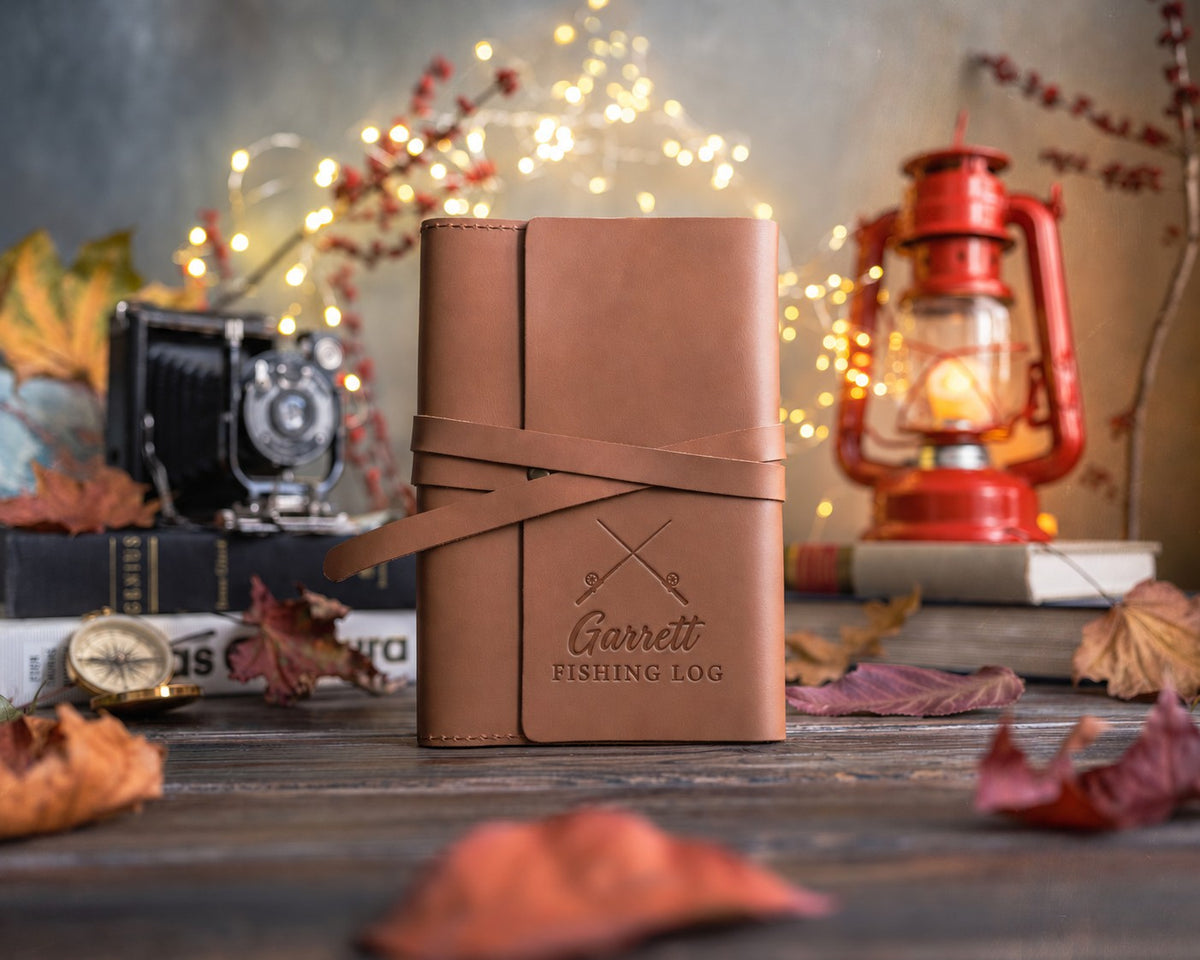 Caramel leather wrap journal with its strap tied, the lower cover debossed with crossed fishing rods above Garrett and FISHING LOG
