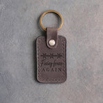 Country gray leather keychain debossed with a band of barbed wire above Fixing fence and AGAIN, on gray plaster