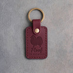 Sangria red leather keychain debossed with a Pomeranian silhouette and the words Floof IN CHARGE, on gray concrete