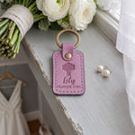 Lily FLOWER GIRL keychain in lilac leather on a gray windowsill beside bouquet stems wrapped in linen