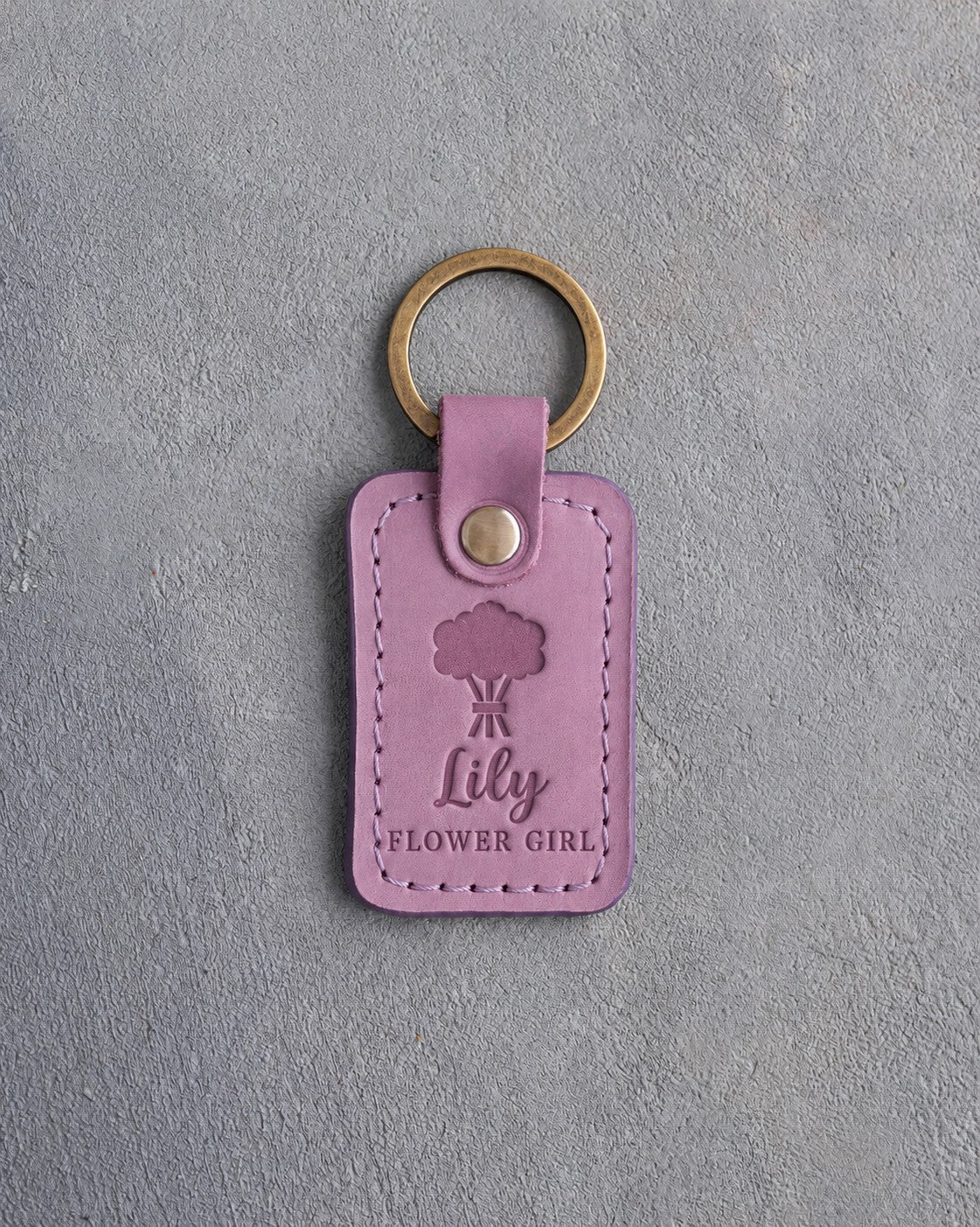 Lilac leather keychain debossed with a hand-tied bouquet above the name Lily and FLOWER GIRL, on gray concrete