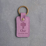 Lilac leather keychain debossed with a hand-tied bouquet above Our and FLOWER GIRL, on gray concrete