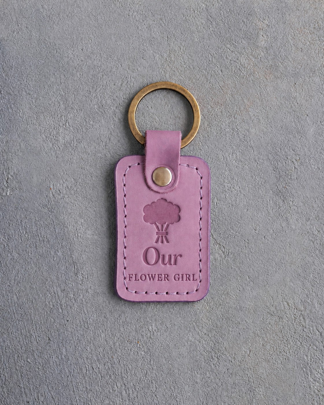 Lilac leather keychain debossed with a hand-tied bouquet above Our and FLOWER GIRL, on gray concrete