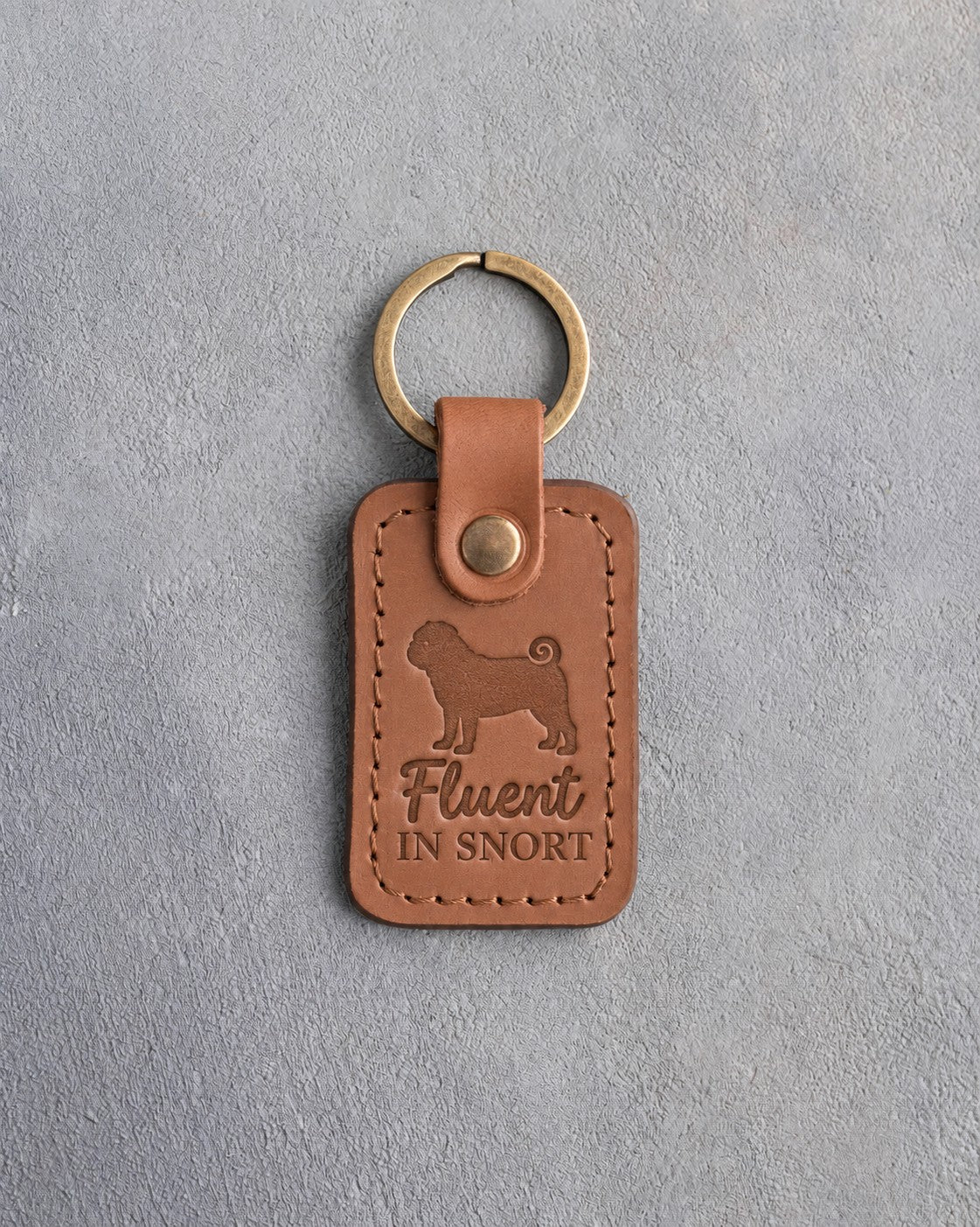 Caramel leather keychain debossed with a pug silhouette and the words Fluent in SNORT, on gray concrete