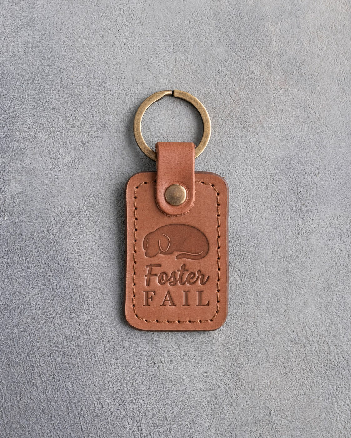 Caramel leather keychain debossed with a curled sleeping dog and the words Foster FAIL, on gray concrete