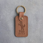 Caramel leather keychain debossed with a goldendoodle standing on its hind legs and the words Free HUGS, on gray concrete